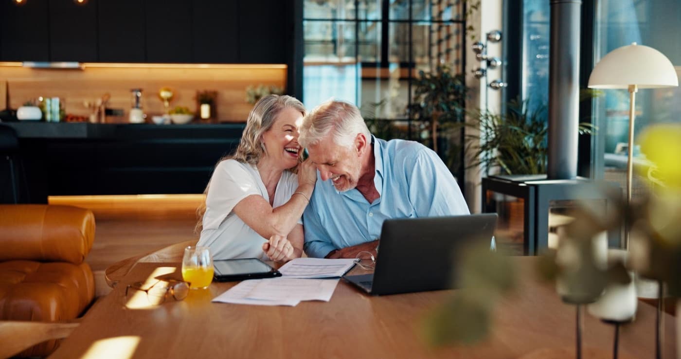 Retired couple laughing while reviewing retirement planning documents on a laptop.