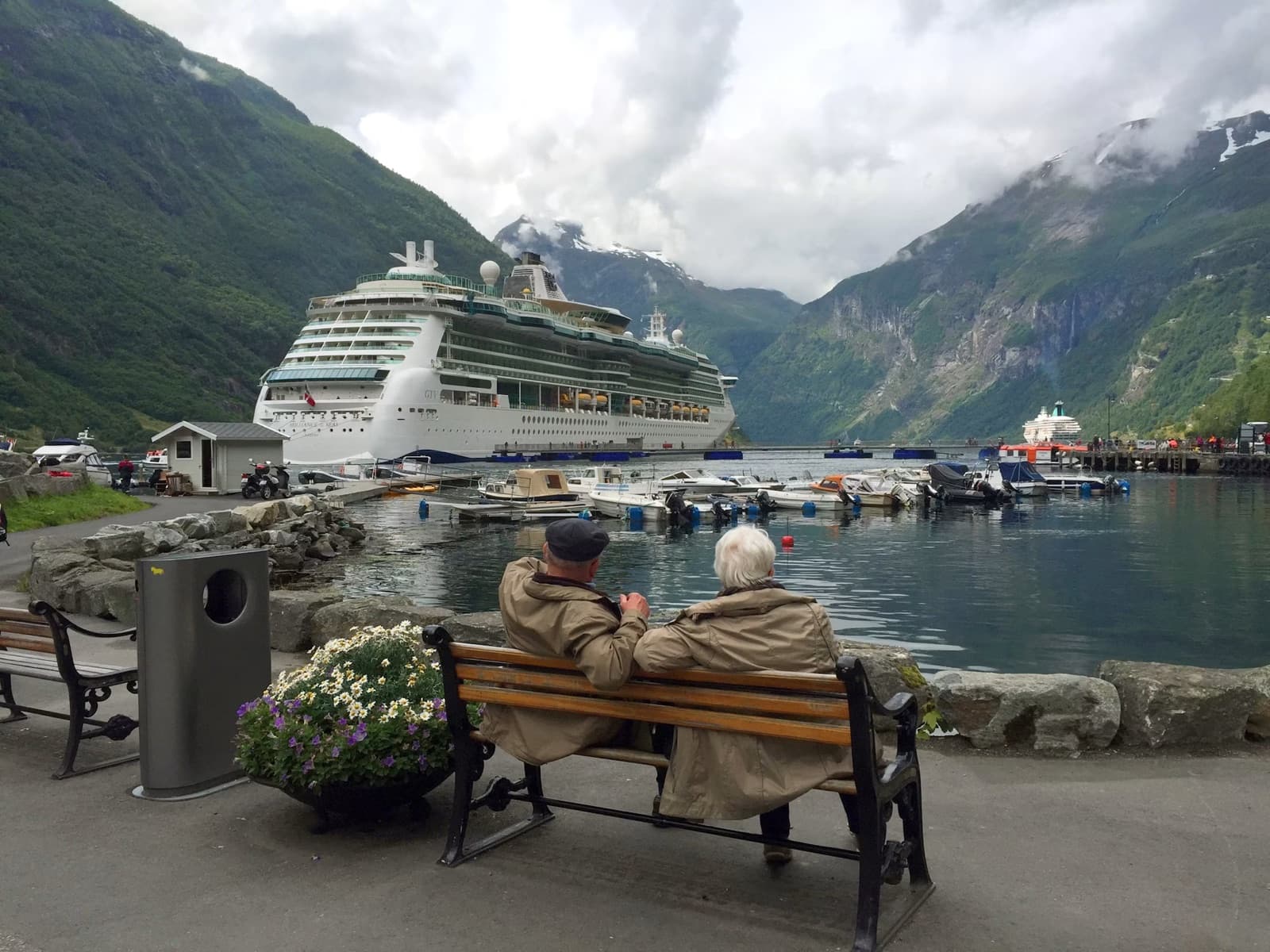 Retired couple sitting by the water near a cruise ship and mountains.